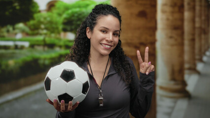 Woman holding soccer ball and showing peace sign in park with lush greenery and stone columns in background, radiating happiness and enthusiasm outdoors.