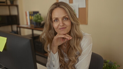 Woman with glasses in office with blonde hair, wearing shirt, sitting indoors, relaxed and thoughtful at workplace setting, modern interior visible with shelves and plants.