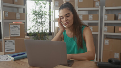 Woman typing on laptop beside stacked cardboard parcels with shipping labels in office; work...