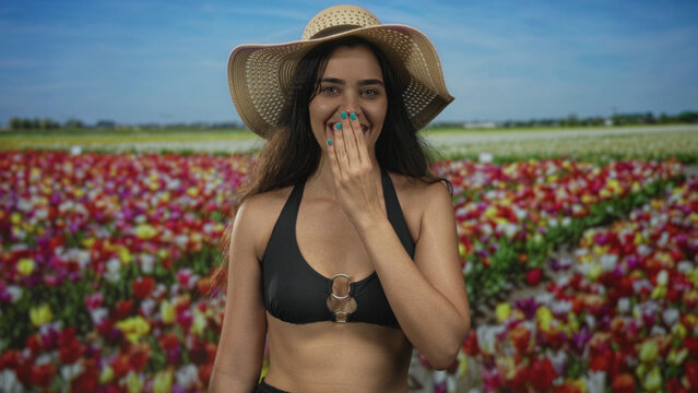 Woman in black bikini top and straw hat covers mouth with hand in forest; joy surprise wonder curiosity.
