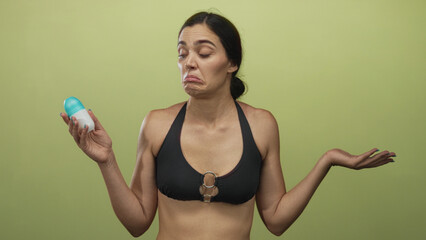 Woman holds roll on deodorant and shrugs with uncertain expression in olive green studio; personal hygiene dilemma.