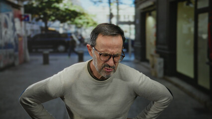 Fototapeta premium Hispanic man with glasses stands outdoors in an urban city street setting, expressing discomfort as he clutches his stomach in mid-day light.