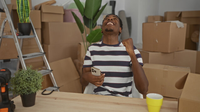 Young man sitting at wooden table holding smartphone at home amid moving boxes clutching chest; stress heartattack.