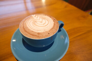 Close-up of a creamy cappuccino topped with beautiful latte art, served in a blue ceramic cup and saucer on a warm wooden surface.  

