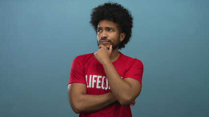 African american lifeguard man in red shirt pensively poses against an isolated blue wall background, embodying thoughtfulness and contemplation.