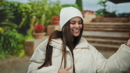 Young hispanic woman smiles while mimicking playing guitar outdoors on a street, wearing a white...