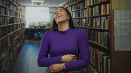 Woman laughs and holds her stomach amid tall library shelves in a quiet aisle wearing a purple...