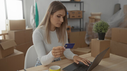 Woman using credit card on laptop in new home surrounded by cardboard boxes with a plant on the table, capturing a moment of online shopping or banking in a fresh setting.