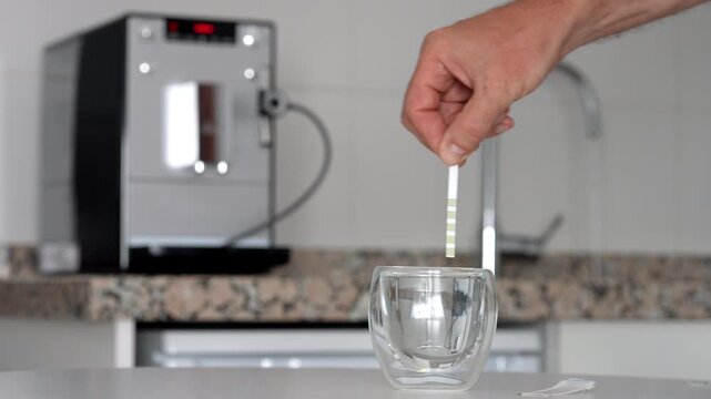 Testing tap water hardness with strip near coffee machine, result shows hard water. Strip dipped for 1 second, shaken, result read after 1 minute.