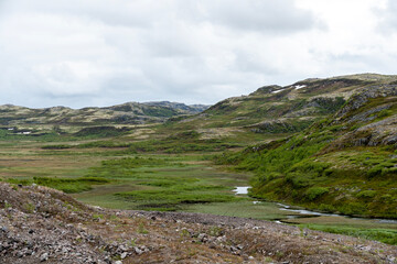 Rolling hills adorned with patches of snow contrast with vibrant green vegetation. The overcast sky adds a mystical quality to the tranquil landscape, creating a peaceful atmosphere.