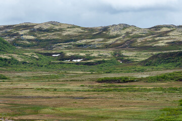 Obraz premium Rolling hills adorned with patches of snow contrast with vibrant green vegetation. The overcast sky adds a mystical quality to the tranquil landscape, creating a peaceful atmosphere.