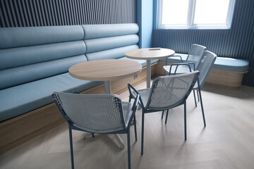 Contemporary lounge space featuring round wooden tables, minimalist chairs, padded blue bench seating, and soft natural lighting from windows.
