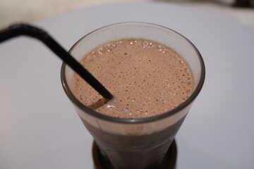 Chilled chocolate beverage served in a clear glass with a black straw, placed on a round table in a minimal indoor setting.
