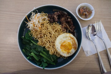 Top-down view of dry instant noodles served with stir-fried beef, fried egg, leafy greens, bean sprouts, and sambal in a dark bowl.
