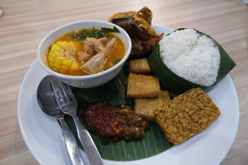 Traditional Indonesian meal featuring white rice, tempeh, tofu, fried chicken, spicy sambal, and sayur asem vegetable soup on a banana leaf.