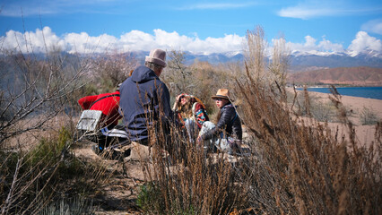 A group of people is sitting on the sand in an overgrown field, with snow-capped mountains in the background. Friends are relaxing in nature