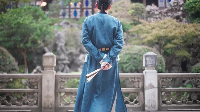 A man in traditional blue Hanfu stands with his back facing the camera holding a fan on a stone bridge in a classical Chinese garden