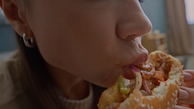 Closeup slowmo shot of young Caucasian woman biting saucy burger enjoying fast food at home