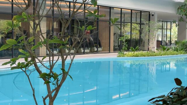 Modern hotel swimming pool near gym with glass wall and tropical plants, tranquil luxury resort atmosphere.