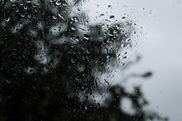 A photo of rain drops on the window glass with a blurred view of the blossoming green trees. Abstract image showing cloudy and rainy weather conditions