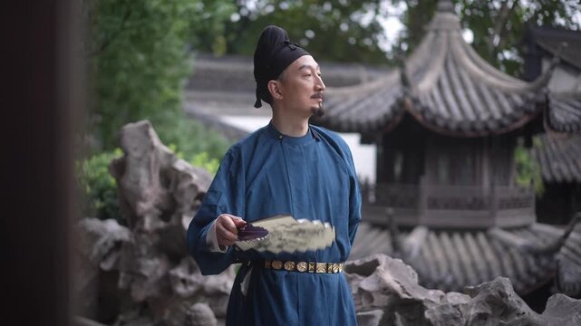 A man in traditional Chinese attire holding a tray stands before rockery and pavilion in classical garden