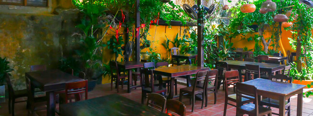 Panorama view Hoi An cafe with red-tiled floor, yellow weathered walls, hanging plants, and rattan lights. Wooden tables and ceiling fans complete this tranquil, garden-style dining space