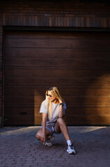 Naklejka premium Stylish young person poses near garage. A young person squats casually on a brick path beside a wooden garage, enjoying the warm sunlight.