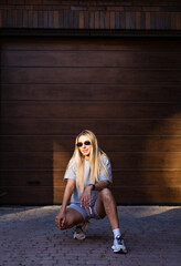 Naklejka premium Stylish young woman outdoors. A woman with long hair squats on the pavement, wearing sunglasses and a casual outfit, enjoying a sunny moment.
