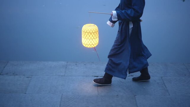 A person in traditional attire walks at night holding a glowing lantern against a cool blue background