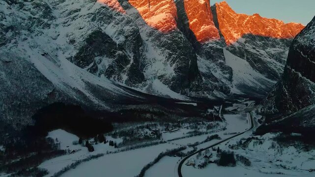 aerial footage capturing the iconic Troll Wall Trollveggen bathed in the warm glow of a winter sunset. Snow covered peaks and dramatic cliffs stand tall in the Romsdalen valley, showcasing the raw bea