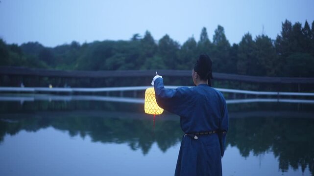 A man in traditional Chinese attire holding a lantern by the lakeside at dusk with reflections on water