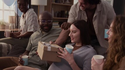 Pregnant woman receiving gift box from friend, opening it and expressing joy while relaxing on couch with young people holding party cups with drinks during baby shower