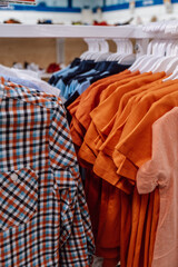 variety brightly colored shirts hang on racks drawn to orange tones and plaid pattern. casual bright clothes for children, simple and comfortable, in baby store. sale. close up.