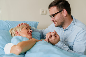 Obraz premium Affectionate Young Man In Glasses Comforting Elderly Mother Lying In Hospital Bed During Family Visit