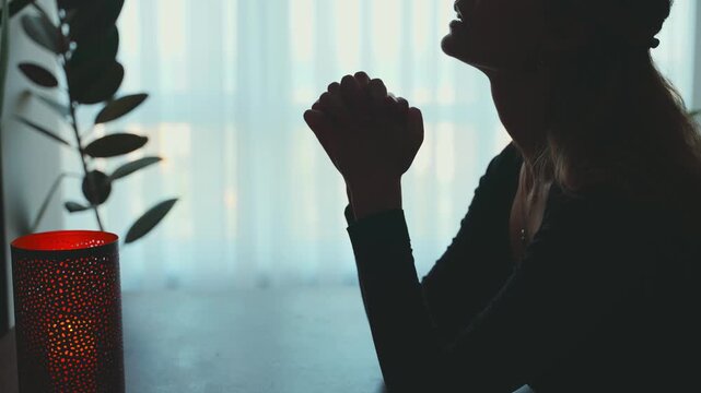 Portrait adult woman praying whispers words of gratitude, hands folded in prayer. Dark silhouette female prayer. Girl Christian praying to God concept of religion faith hope blessing. home room window