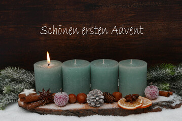 Erster Advent: Natürliche Adventsdekoration zum ersten Advent mit grünen Kerzen, Zapfen und Tannenzweigen im Schnee.   Auf dem hölzernem Hintergrund steht der Gruß einen schönen ersten Advent.