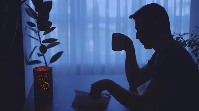 dark silhouette man close up hand flip through paper pages enjoy reading book near window dusk blue light home room candle. guy sitting at desk hand holding cup tea coffee hot drink. hobby rest relax