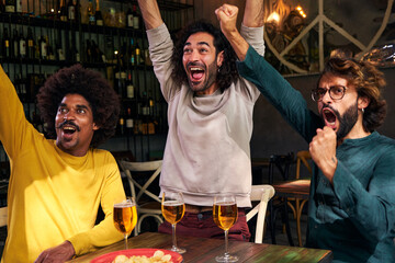 Multiethnic group of three male friends watching soccer match at a bar. Celebrating happily a score with beers and chips, gesturing fists up.