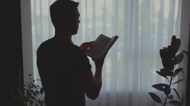unrecognizable person dark silhouette young man hands fingers flip through paper pages reading book near window dusk light white curtains home black shadow room. Adult guy enjoying hobby rest relax