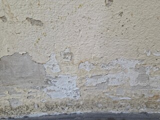 A damaged wall with peeling yellowish paint on the upper section, exposing grey concrete or plaster below, showing flaking, discoloration, and possible mold or dirt.