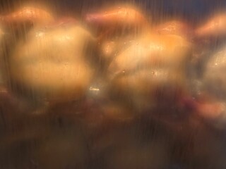 Several whole chickens roasting inside a rotisserie machine, seen through the fogged or steam-covered glass while they cook, give the scene a blurred and hazy appearance.
