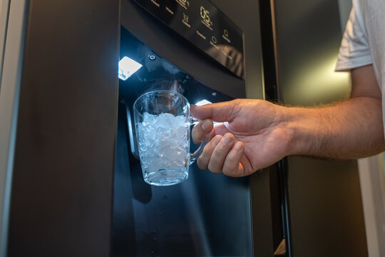 Hand holds a glass under a refrigerator ice cube dispenser. The cup is filled with fresh crushed ice.