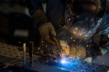 Man with welding mask in safety suit with gloves welds steel in home workshop. Welding flame with sparks. Dangerous work