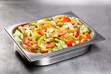 A colorful garden salad filled with crisp lettuce, sliced tomatoes, and vibrant green peppers is displayed in a stainless steel container. The kitchen is busy, indicating food preparation activity