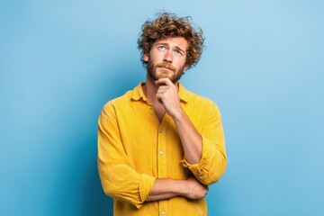 Confident young man pondering thoughtfully against a solid blue wall while wearing a casual bright yellow shirt, showcasing a relaxed and introspective attitude during the daytime