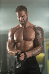 Fototapeta premium muscular man stands in a gym tightening straps on his arm in preparation for a weightlifting workout. The atmosphere is filled with fitness equipment and a focus on strength training.