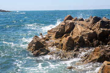 Rocky coastline with waves crashing against jagged rocks under a clear sky, vibrant ocean water and sea spray