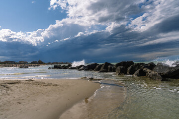 Beach scene with dark rocks, waves crashing, dramatic sky, and sand in the foreground under cloudy sky. Serene yet powerful coastal view.