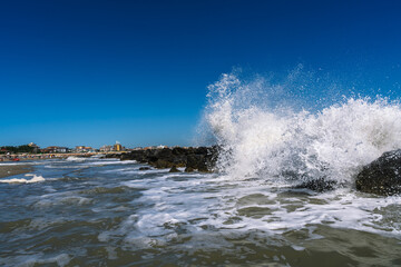Waves crashing against rocks under a clear blue sky near a coastal town, capturing the ocean's raw power and scenic beauty.