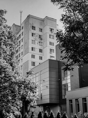Monochrome view of two buildings with contrasting styles, a weathered high-rise and a modern, glass-fronted structure framed by dense foliage